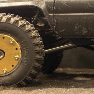 May include: Close-up of a black toy truck with a large, knobby tire. The tire has a gold-colored center with multiple bolts. The truck's body is black and shows signs of wear.