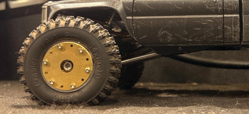 May include: Close-up of a black toy truck with a large, knobby tire. The tire has a gold-colored center with multiple bolts. The truck's body is black and shows signs of wear.