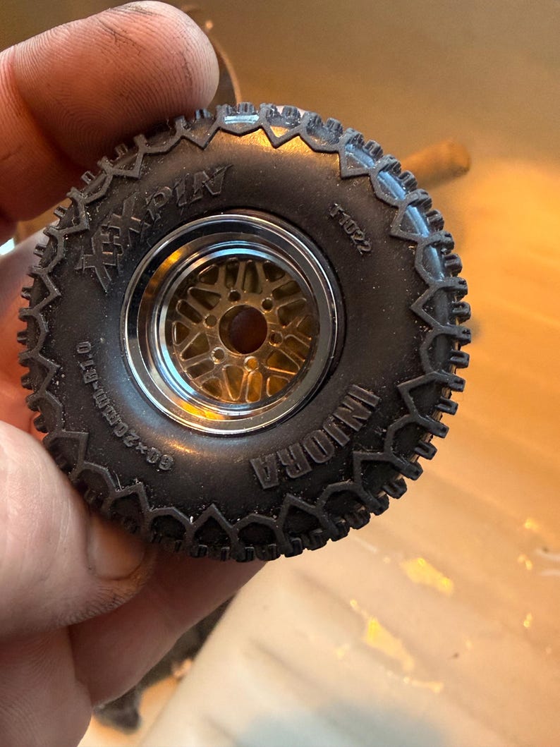 May include: A black rubber tire with a deep tread pattern mounted on a gold-colored metal wheel with a chrome rim. The tire sidewall features the text INJORA, XXPIN, T1022, and 60x20mm-R1.0 molded into the surface of the rubber material.