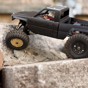 May include: A matte black remote-controlled pickup truck model with a textured body, featuring a gold-colored front wheel rim, black rear wheel rims, and visible internal electronic components through the open cabin and truck bed area.