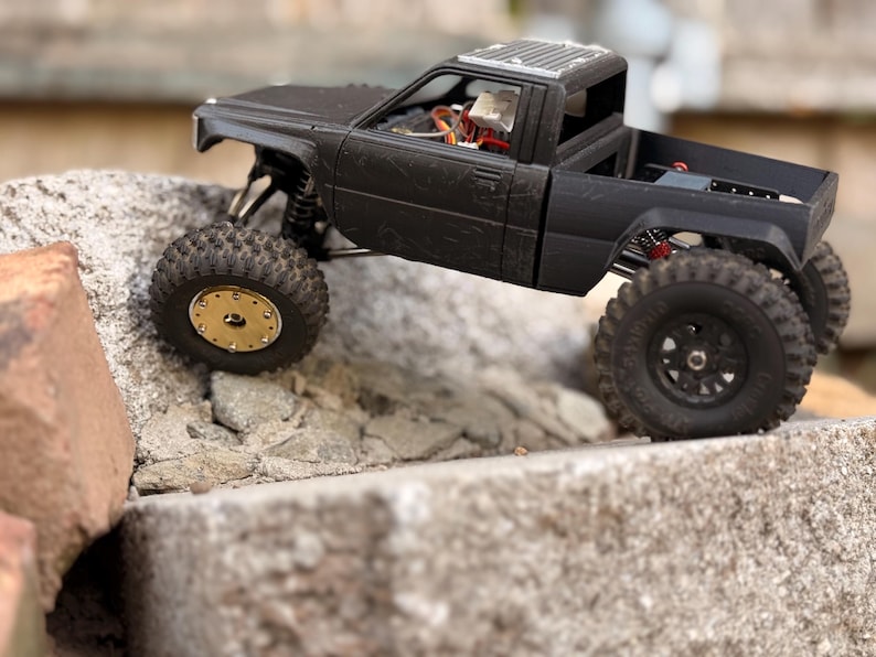 May include: A matte black remote-controlled pickup truck model with a textured body, featuring a gold-colored front wheel rim, black rear wheel rims, and visible internal electronic components through the open cabin and truck bed area.