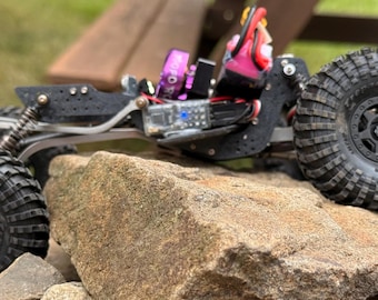 May include: A remote-controlled rock crawler vehicle featuring large black knobby tires, a black chassis, exposed mechanical suspension components, and a purple motor component, positioned on top of a cluster of jagged brown rocks.