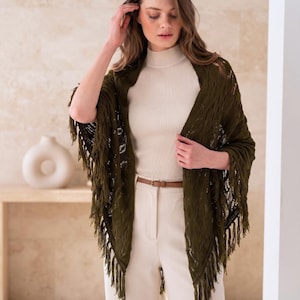 May include: An olive green triangular shawl with an open-knit pattern and long fringe edges, worn over a cream-colored mock-neck top and light-colored trousers with a thin brown belt. The logo "aas adaapparelstudio" is in the top left corner.