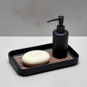 May include: A black soap dispenser and a bar of soap rest on a black tray with a wooden slat insert. The soap dispenser has a black pump. The oval soap bar is light beige with the word "CREATE" embossed on it.