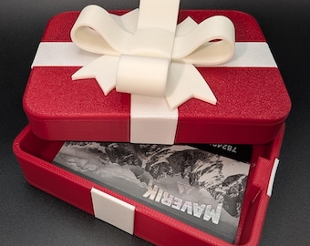 Può includere: Una scatola regalo rossa con fiocco e nastro bianchi. La scatola è aperta, rivelando una carta con la scritta "MAVERIK" stampata sopra. La scatola è di forma quadrata.
