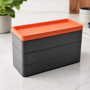 May include: A rectangular, three-tiered storage container featuring two dark gray base sections and a vibrant orange lid. The stackable unit has a smooth, matte finish and clean, straight edges, providing a structured appearance for organization.