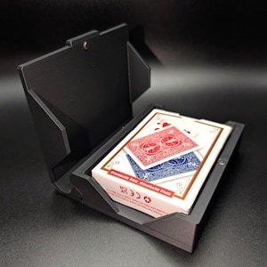 May include: A black, geometric playing card case with a deck of cards inside. The cards are red and blue with a white border. The case is open, revealing the cards. The case is made of a matte black material.