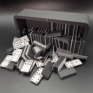 May include: A gray domino set with black and white tiles spilling out of a matching gray case. The dominoes are black with white dots, and the case is open, revealing neatly stacked dominoes inside.