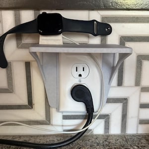 May include: A black smartwatch rests on a white charging stand above an electrical outlet. The stand is mounted on the wall and has a shelf. A black power cord is plugged into the outlet.