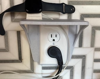 May include: A black smartwatch rests on a white charging stand above an electrical outlet. The stand is mounted on the wall and has a shelf. A black power cord is plugged into the outlet.