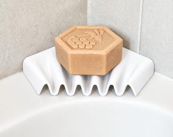May include: A white, wavy-edged corner soap dish holding a hexagonal tan bar of soap. The soap features a raised, embossed design of a bee resting on a honeycomb pattern on its top surface, centered within the dish.