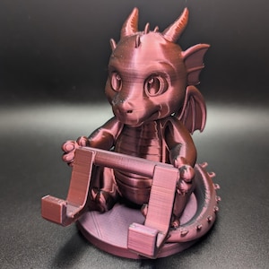 May include: A maroon-colored dragon figurine with a phone or tablet stand. The dragon has large eyes, horns, wings, and a spiky tail. The stand is integrated into the design, held by the dragon's hands.