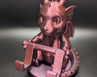 May include: A maroon-colored dragon figurine with a phone or tablet stand. The dragon has large eyes, horns, wings, and a spiky tail. The stand is integrated into the design, held by the dragon's hands.