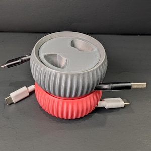 May include: A two-piece cable organizer in gray and red. The top gray piece has a lid with three openings. The bottom red piece has a ribbed design. Cables with black and white connectors are plugged into the organizer.