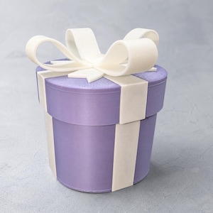 May include: A cylindrical gift box in a light purple shade with a smooth finish. The box features a wide, matte white ribbon that wraps vertically around the sides and lid, topped with a large, decorative white bow made of looped ribbon.