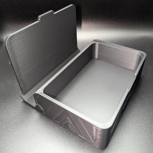 May include: A dark gray, rectangular box with a hinged lid, open to reveal the interior. The box has a textured surface and geometric design elements on the side. The lid is partially open, showing the inside of the box.