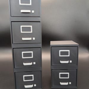 May include: Two sets of miniature black filing cabinets. One set is stacked with five drawers, and the other has two drawers. Each drawer has a white rectangular label holder and a small white handle.