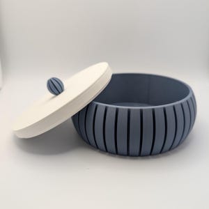 May include: A blue and white decorative storage bowl with a lid. The bowl is a light blue color with vertical grooves. The lid is white with a small blue sphere on top. The bowl is open, revealing the interior.