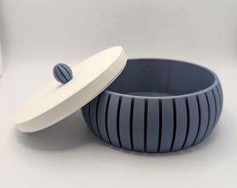 May include: A blue and white decorative storage bowl with a lid. The bowl is a light blue color with vertical grooves. The lid is white with a small blue sphere on top. The bowl is open, revealing the interior.