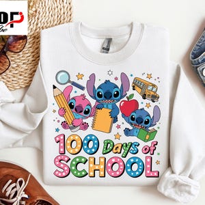 May include: White sweatshirt with a colourful cartoon design and the text "100 Days of School." The design includes a pink character holding a pencil, blue characters with a notebook and book, a red apple, and a yellow school bus.