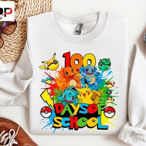 Op de afbeelding: Witte sweatshirt met kleurrijke cartoonfiguren en de tekst "100 DAYS OF SCHOOL". Het ontwerp bevat afbeeldingen van verschillende geanimeerde personages met een splash van verf op de achtergrond. Een leuk en levendig ontwerp.
