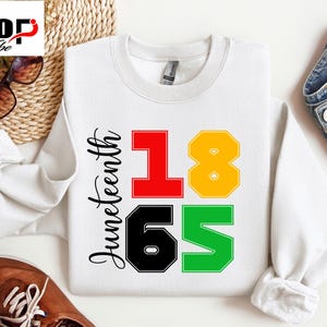 Può includere: Felpa bianca con la scritta "Juneteenth" in corsivo nero. I numeri "1865" sono in rosso, giallo, nero e verde. Il logo SVG Pop Vibe è nell'angolo in alto a sinistra. Occhiali da sole e un cestino intrecciato sullo sfondo.