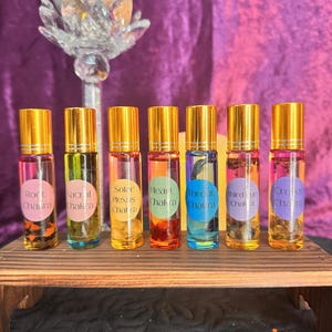 May include: A collection of seven glass roller bottles with gold caps, each filled with a different colored liquid. The bottles are labeled with the names of chakras: Root, Sacral, Solar Plexus, Heart, Throat, Third Eye, and Crown. The bottles are displayed on a small wooden shelf.