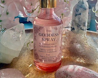 Może przedstawiać: Różowa szklana butelka mgiełki rytualnej "The Goddess Spray" z różą, ze złotą nakrętką, stoi wśród kryształów i różowych kwiatów. Etykieta głosi "Infused with Rose, Quartz & Sandalwood" i "2 oz".