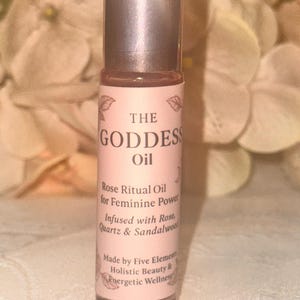 May include: A small, clear glass roll-on bottle with a silver cap. The label is light pink with the words "THE GODDESS Oil" and other text. The oil is for feminine power and infused with rose, quartz, and sandalwood.