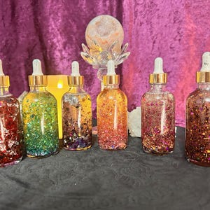 May include: Six glass dropper bottles filled with colorful glitter and liquid. The bottles have gold-colored caps and are arranged on a black surface. A lit candle and a crystal sphere are in the background, against a purple backdrop.