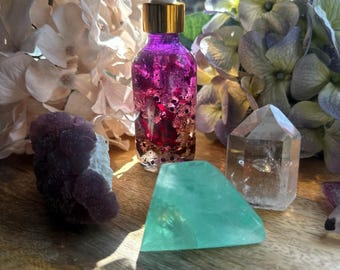 May include: A purple and gold essential oil bottle with a dropper, surrounded by crystals and flowers. The bottle contains a pink liquid with gold flecks. A green fluorite crystal and a clear quartz crystal are also visible.