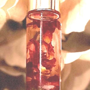 May include: A clear glass roll-on bottle with a silver cap, filled with a reddish-brown liquid and flower petals. The bottle sits on a light-colored, textured surface, with a soft, blurred background.