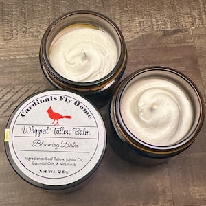 May include: Three small jars of whipped tallow balm. The jars have a dark blue and gold rim. The label reads "Cardinals Fly Home Whipped Tallow Balm Blooming Balm" with ingredients listed. Each jar contains 2 oz of product.