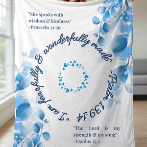 Puede incluir: Manta blanca con diseño floral y de hojas azules. La manta tiene varios versículos bíblicos impresos, incluyendo "Ella habla con sabiduría y bondad" y "El Señor es mi fuerza y mi canción."