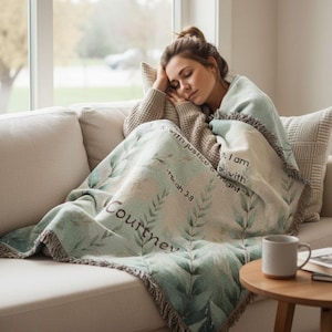 May include: A light green and cream-colored throw blanket with a leaf design and the name "Courtney" printed on it. The blanket also features the text "Micah 3:8" and other script. The blanket has a fringed edge.