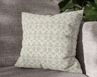 May include: Square decorative pillow with a cream background and a repeating pattern of interconnected gold and teal floral designs. The pillow is placed on a gray sofa, suggesting a home decor theme.