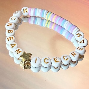 May include: A beaded bracelet with white letter beads spelling "ONBEAM ICECREAM" and a gold star charm. The bracelet features a section of pastel-colored disc beads. The bracelet is reflected on a shiny surface.