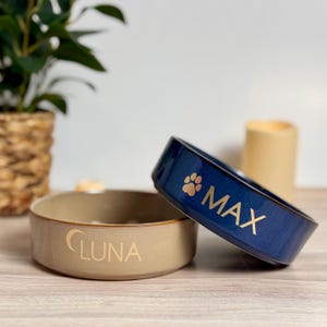 May include: Two ceramic pet bowls. One is beige with "LUNA" in gold lettering. The other is navy blue with "MAX" in gold lettering and a paw print. Both bowls are on a wooden surface.