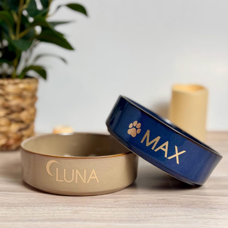 May include: Two ceramic pet bowls. One is beige with "LUNA" in gold lettering. The other is navy blue with "MAX" in gold lettering and a paw print. Both bowls are on a wooden surface.
