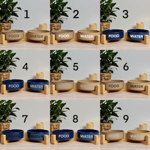 May include: A collection of pet bowls in various colors, including beige and navy blue. Each bowl is labeled with either "FOOD" or "WATER" in a simple font. The bowls are set on wooden stands, with a plant in the background.