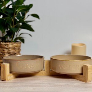 May include: A tan ceramic pet bowl set with a bamboo stand. One bowl is labeled "LUNA", the other "WATER". The bowls are round and sit on a light brown bamboo stand. A green plant and a candle are in the background.