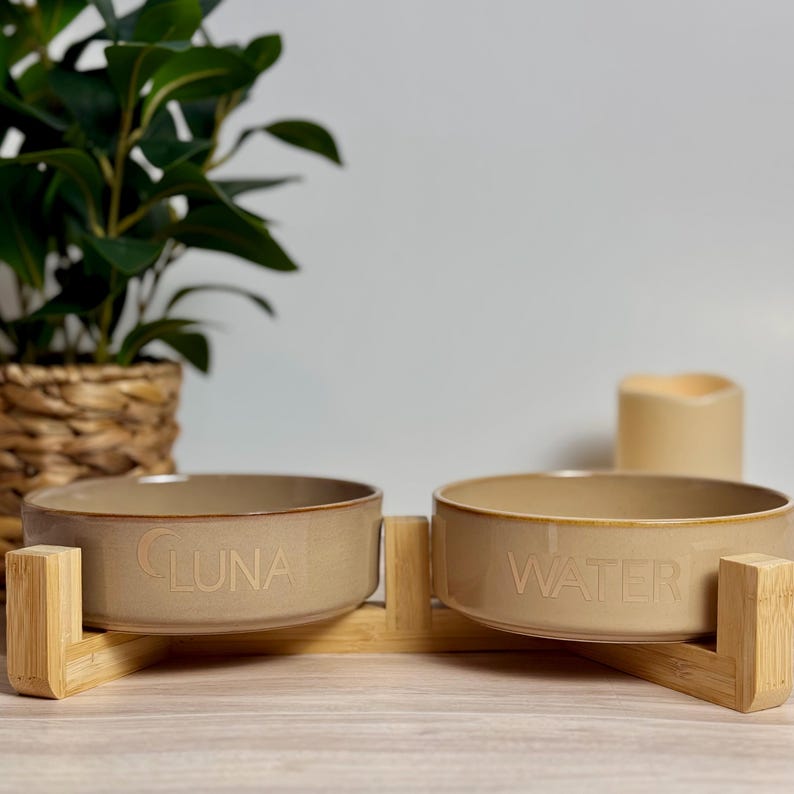 May include: A tan ceramic pet bowl set with a bamboo stand. One bowl is labeled "LUNA", the other "WATER". The bowls are round and sit on a light brown bamboo stand. A green plant and a candle are in the background.