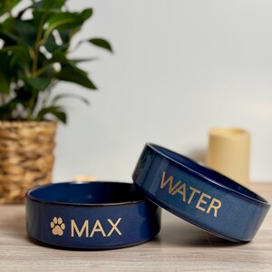 May include: Two navy blue ceramic pet bowls. One bowl has the word "MAX" and a paw print, the other says "WATER" in gold lettering. The bowls are on a wooden surface, with a plant and candle in the background.