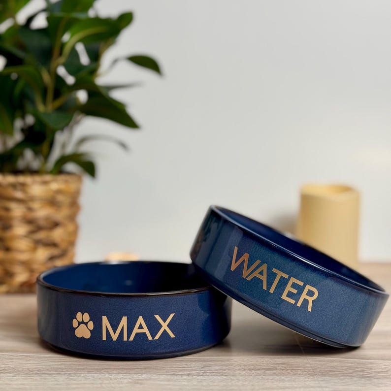 May include: Two navy blue ceramic pet bowls. One bowl has the word "MAX" and a paw print, the other says "WATER" in gold lettering. The bowls are on a wooden surface, with a plant and candle in the background.