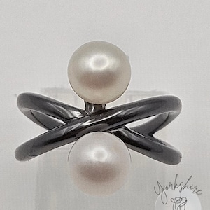 May include: A black metal ring with two white pearls. The ring's band has an X-shaped design, with a pearl at the top and bottom of the X. The pearls are round and have a lustrous finish. The ring is set against a white background.