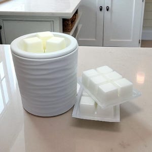 May include: A white ceramic wax warmer with a wavy design, containing four white wax cubes. Two packages of wax cubes are placed next to the warmer. The setting is a bathroom with a neutral colour scheme.
