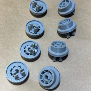 May include: A collection of eight gray, 3D-printed plastic components. Each component has a circular base with a central structure featuring multiple prongs and a raised outer ring. The parts are arranged on a light brown surface.