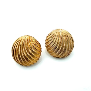 May include: A pair of round, gold-toned metal earrings featuring a textured, spiraling ribbed pattern that radiates from the center. The surface of each earring has a brushed finish with defined, raised ridges that create a shell-like appearance.