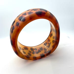 May include: A wide, oval-shaped bangle bracelet with a tortoiseshell pattern. The bracelet features a warm, amber-brown hue with dark brown spots, creating a classic and stylish accessory. The bangle is made of a smooth, glossy material.