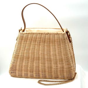 May include: Woven wicker handbag with a brown leather handle and a gold-tone chain strap. The bag has a rectangular shape with a slightly tapered design. The interior is lined with a light-colored fabric.